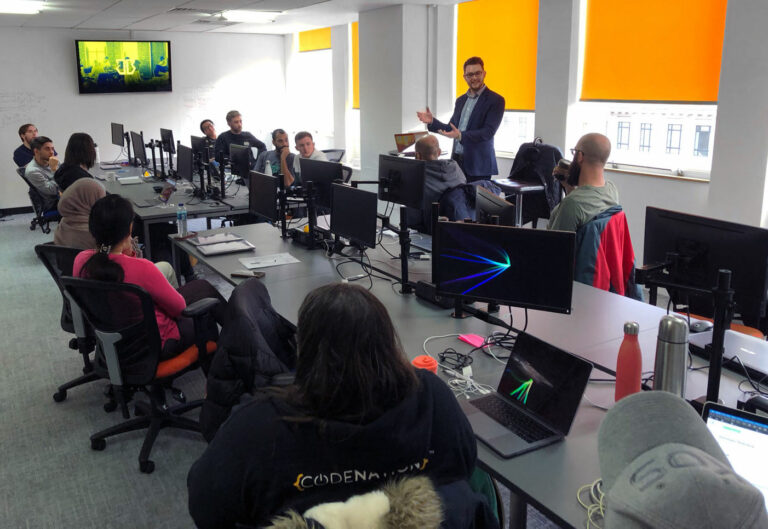 A large group of people are sat at desks listening to someone talk.
